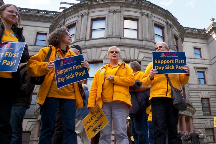 A protest sign reading 'Paid Sick Days Save Lives' held aloft in a crowd of workers